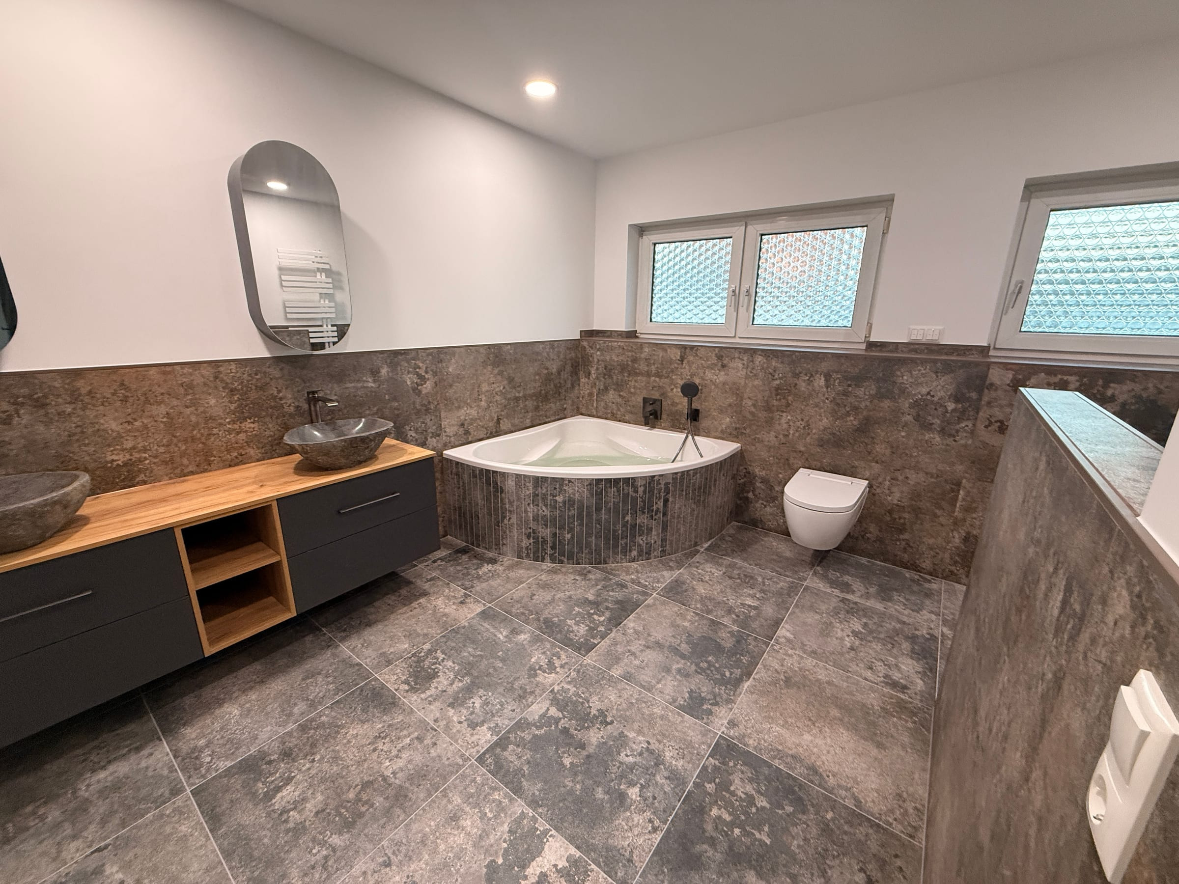 Saniertes Bad mit großformatigen Fliesen in Rostoptik, Eckbadewanne, wandhängendem WC und Waschtisch mit zwei Natursteinbecken.