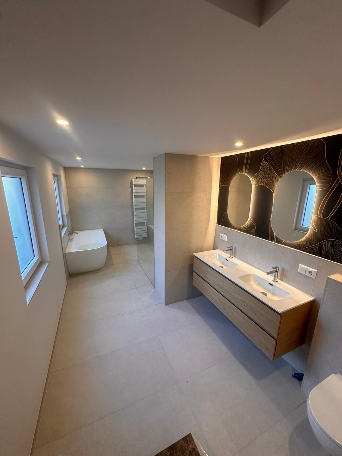 Modernes Master-Bad mit freistehender Badewanne, Walk-in-Dusche, Doppelwaschtisch und beleuchteter Ginkgo-Motivwand mit zwei ovalen Spiegeln.