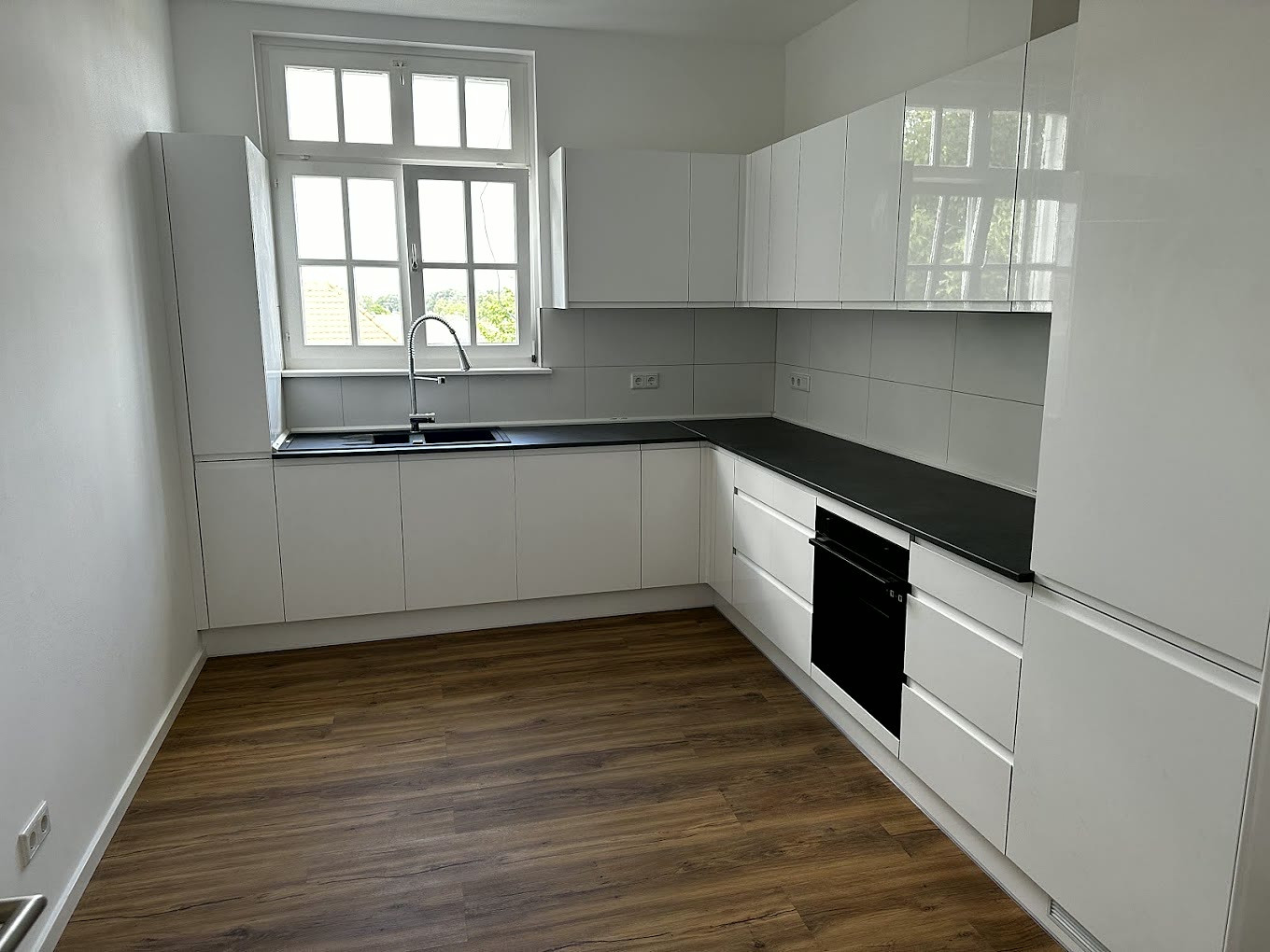 Weiße grifflose Einbauküche mit dunkler Arbeitsplatte, schwarzem Einbaubackofen und Holzboden vor einem Sprossenfenster.